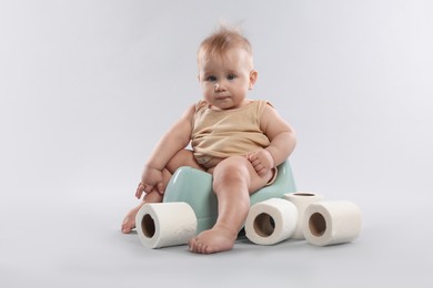 Photo of Cute little baby sitting on potty and rolls of toilet paper against light grey background