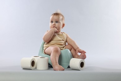 Photo of Cute little baby sitting on potty and rolls of toilet paper against light grey background