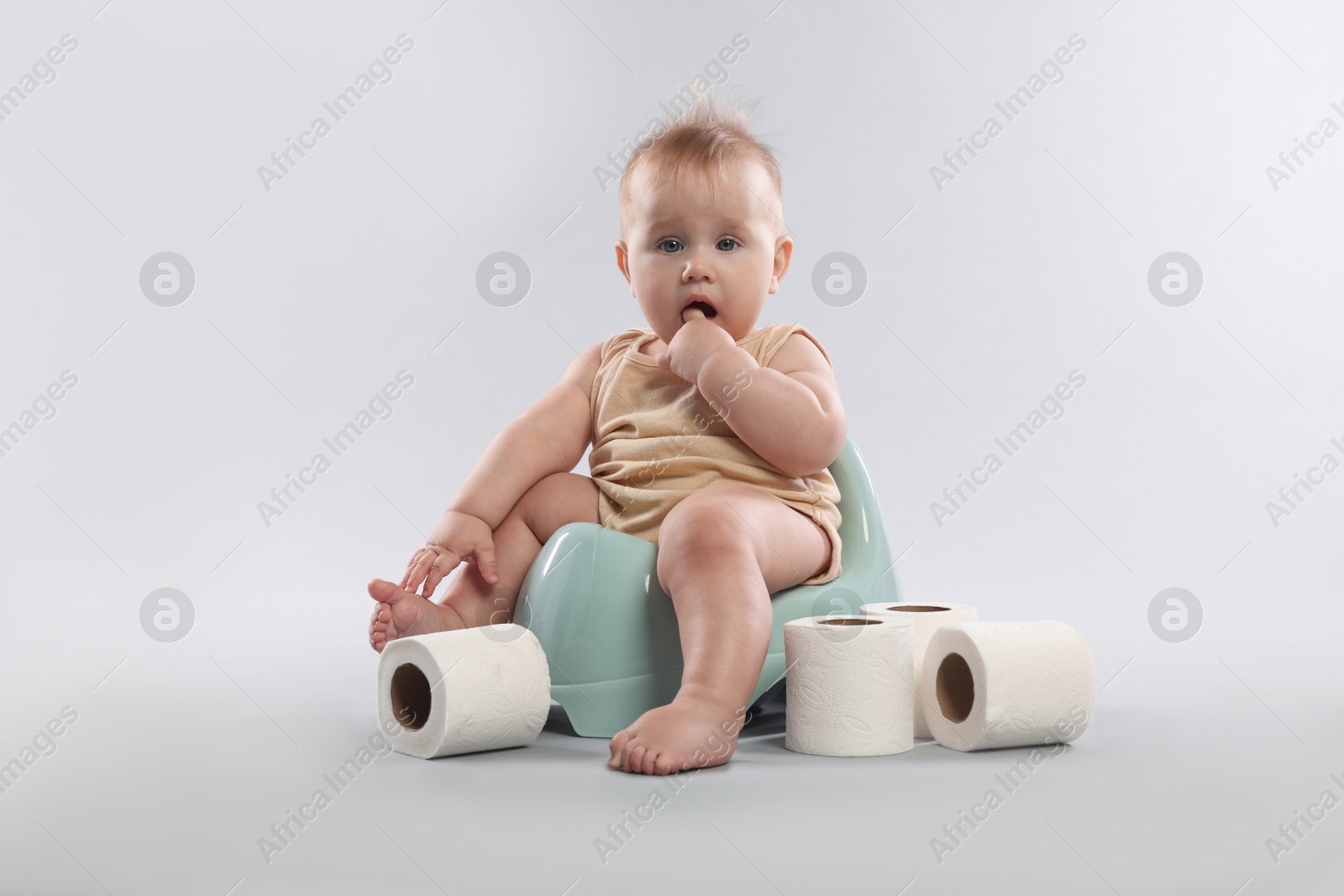Photo of Cute little baby sitting on potty and rolls of toilet paper against light grey background