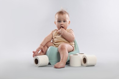 Photo of Cute little baby sitting on potty and rolls of toilet paper against light grey background
