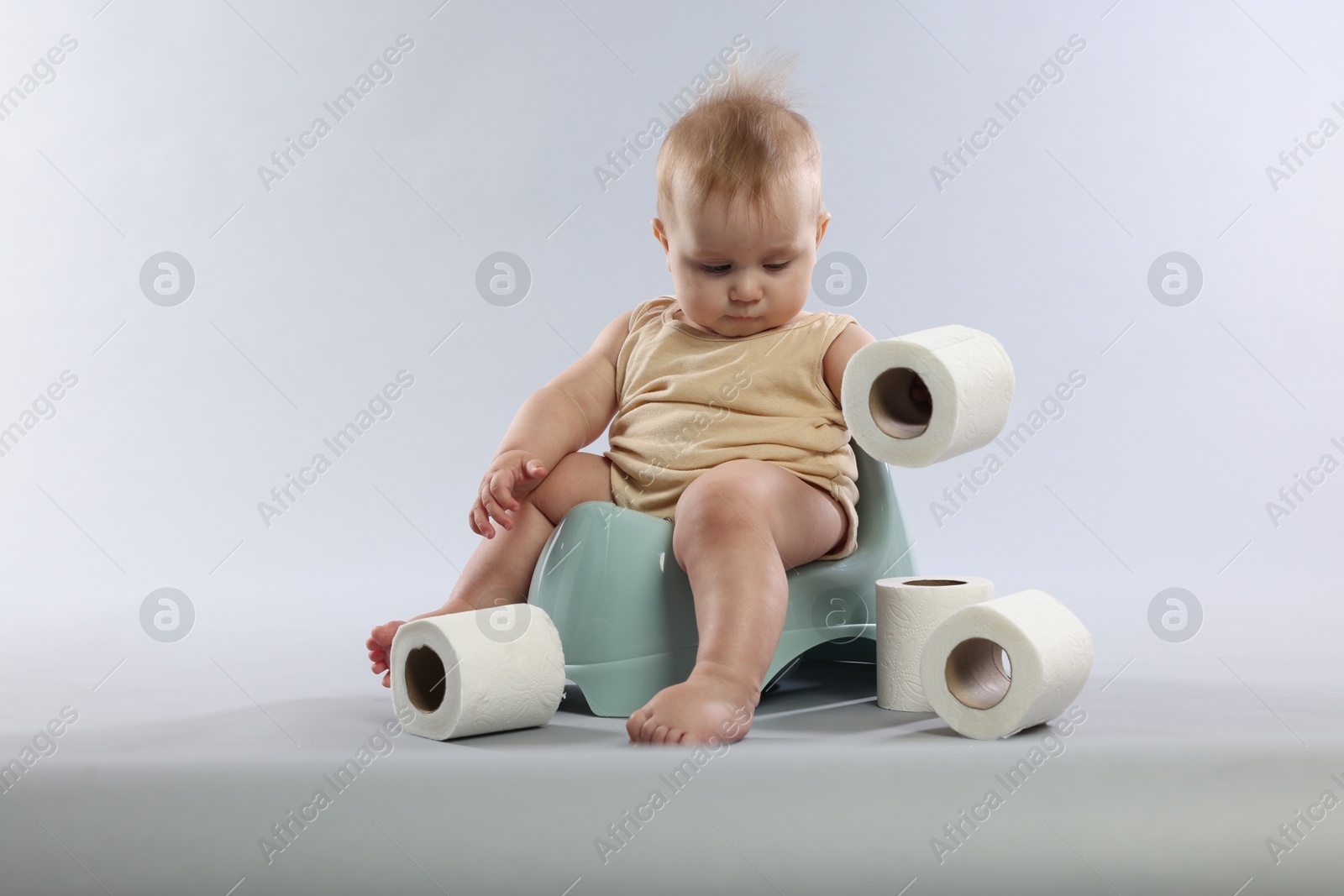Photo of Cute little baby sitting on potty and rolls of toilet paper against light grey background