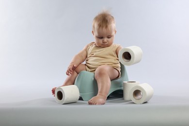 Photo of Cute little baby sitting on potty and rolls of toilet paper against light grey background