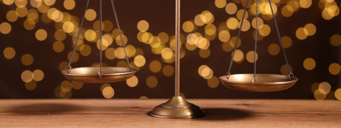 Photo of Golden scales of justice on wooden table against blurred lights, closeup