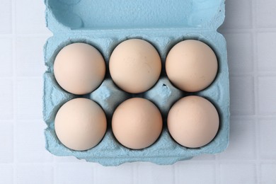 Photo of Raw chicken eggs in egg carton on white tiled table, top view