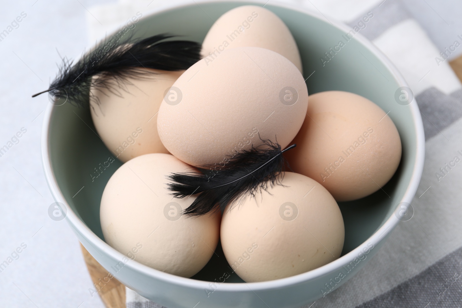 Photo of Raw chicken eggs and feathers in bowl on white table, closeup