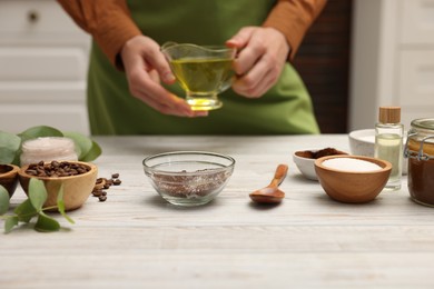 Photo of Making natural scrub. Woman adding oil into bowl at white wooden table, closeup