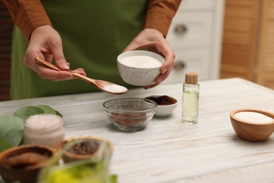 Photo of Making natural scrub. Woman adding sugar into bowl at white wooden table, closeup
