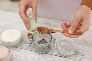 Photo of Making natural scrub. Woman adding essential oil into jar at white wooden table, closeup