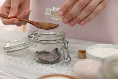Photo of Making natural scrub. Woman adding essential oil into jar at white wooden table, closeup