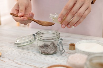 Photo of Making natural scrub. Woman adding essential oil into jar at white wooden table, closeup
