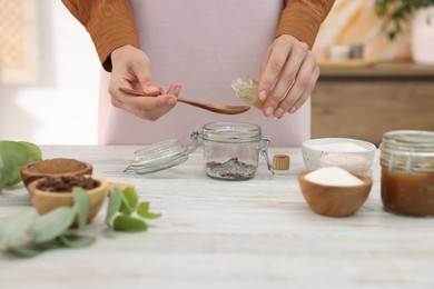 Photo of Making natural scrub. Woman adding essential oil into jar at white wooden table, closeup