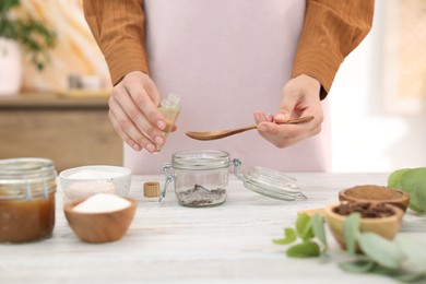 Photo of Making natural scrub. Woman adding essential oil into jar at white wooden table, closeup