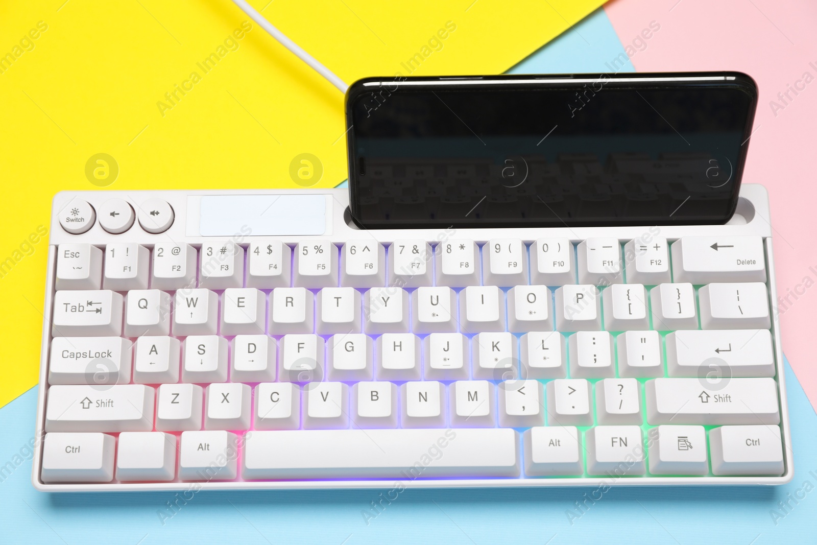 Photo of Modern white keyboard with RGB lighting and smartphone on color background, above view