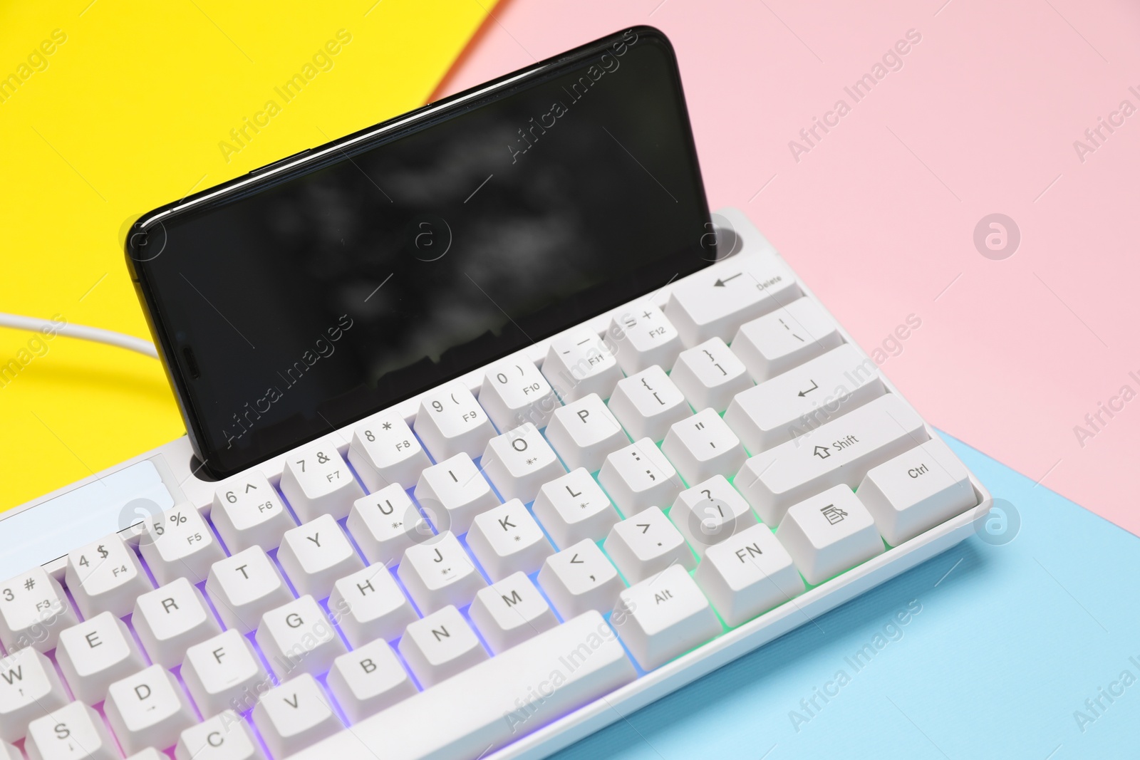 Photo of Modern white keyboard with RGB lighting and smartphone on color background, closeup