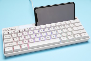 Photo of Modern white keyboard with RGB lighting and smartphone on light blue background, closeup
