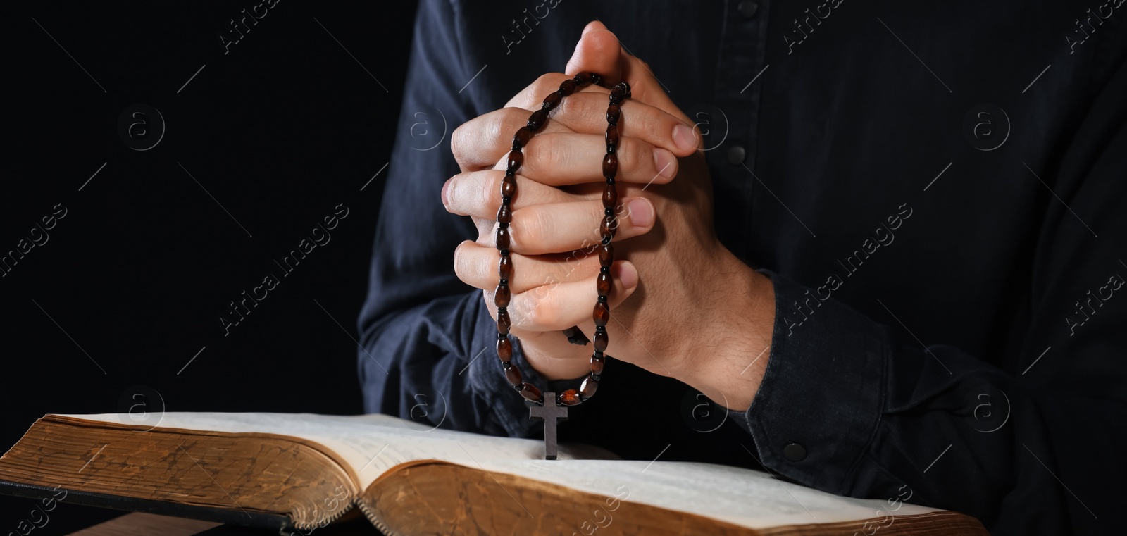 Photo of Priest with rosary and Holy Bible praying on black background, closeup