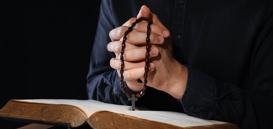 Photo of Priest with rosary and Holy Bible praying on black background, closeup