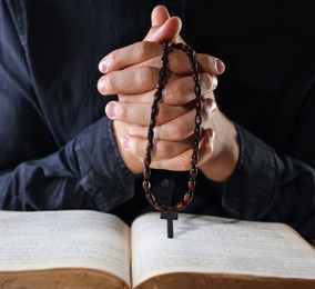 Photo of Priest with rosary and Holy Bible praying on black background, closeup