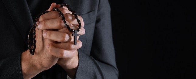 Photo of Priest with rosary praying on black background, closeup. Space for text