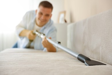 Photo of Man cleaning sofa with vacuum cleaner at home, selective focus