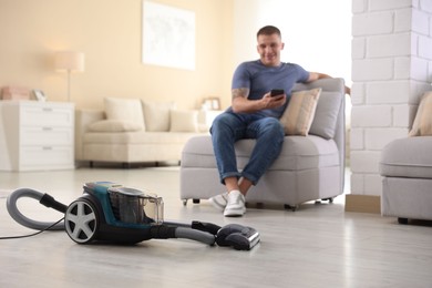 Photo of Man with smartphone on sofa at home, focus on vacuum cleaner