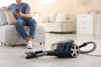 Photo of Man with smartphone on sofa at home, focus on vacuum cleaner