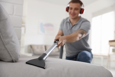 Photo of Man in headphones cleaning sofa with vacuum cleaner at home, selective focus