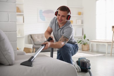 Photo of Man in headphones cleaning sofa with vacuum cleaner at home