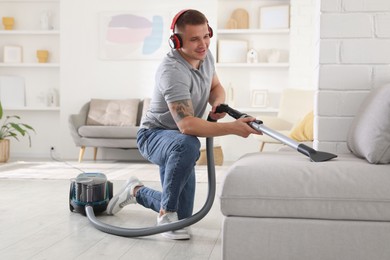 Photo of Man in headphones cleaning sofa with vacuum cleaner at home