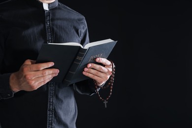Photo of Priest with rosary and Holy Bible on black background, closeup. Space for text