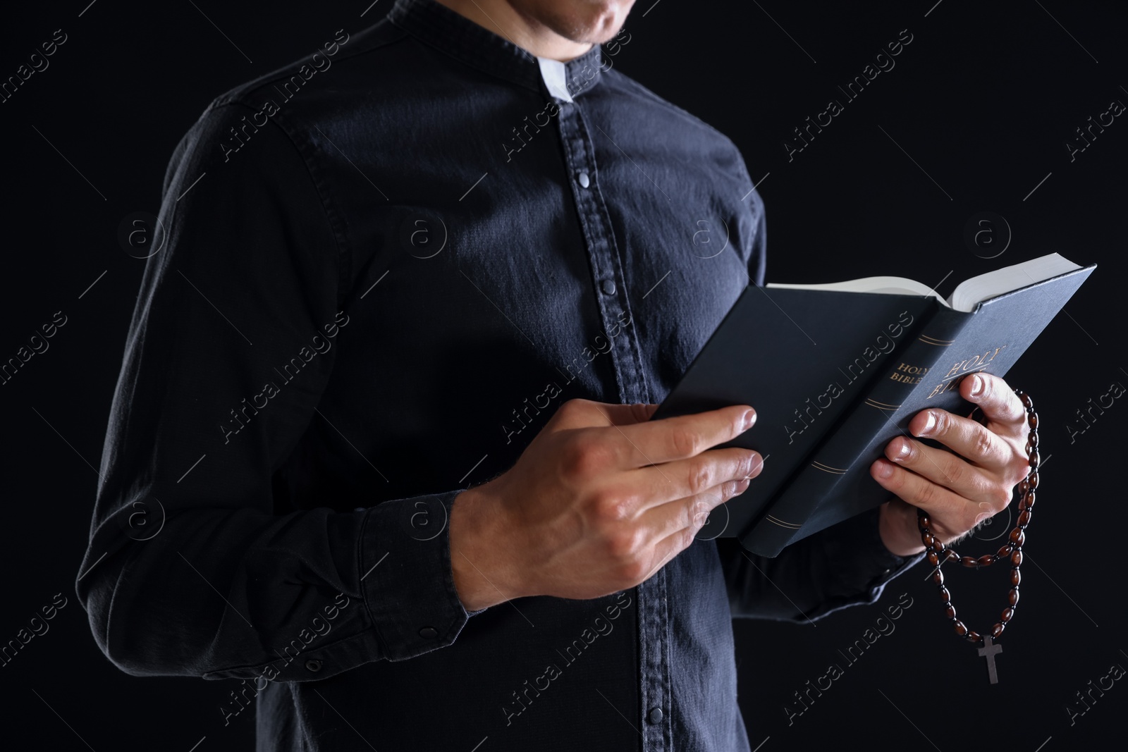 Photo of Priest with rosary and Holy Bible on black background, closeup