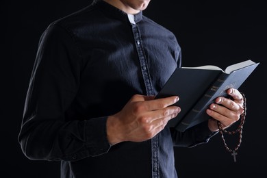 Photo of Priest with rosary and Holy Bible on black background, closeup