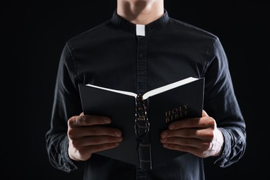 Photo of Priest with Holy Bible on black background, closeup