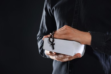 Photo of Priest with rosary and Holy Bible on black background, closeup. Space for text