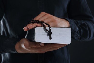 Photo of Priest with rosary and Holy Bible on black background, closeup