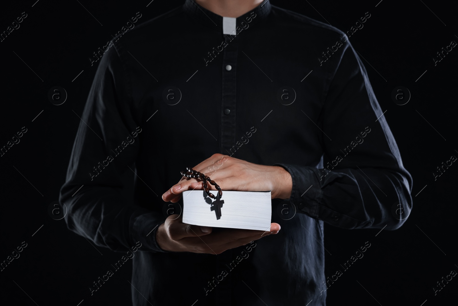 Photo of Priest with rosary and Holy Bible on black background, closeup