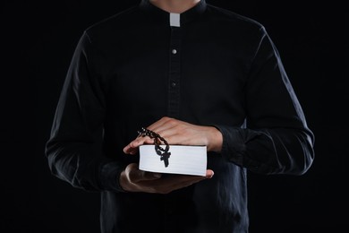 Photo of Priest with rosary and Holy Bible on black background, closeup