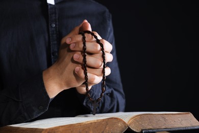 Photo of Priest with rosary and Holy Bible praying on black background, closeup. Space for text