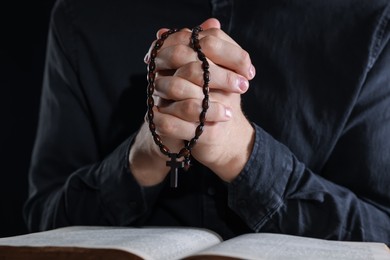 Photo of Priest with rosary and Holy Bible praying on black background, closeup
