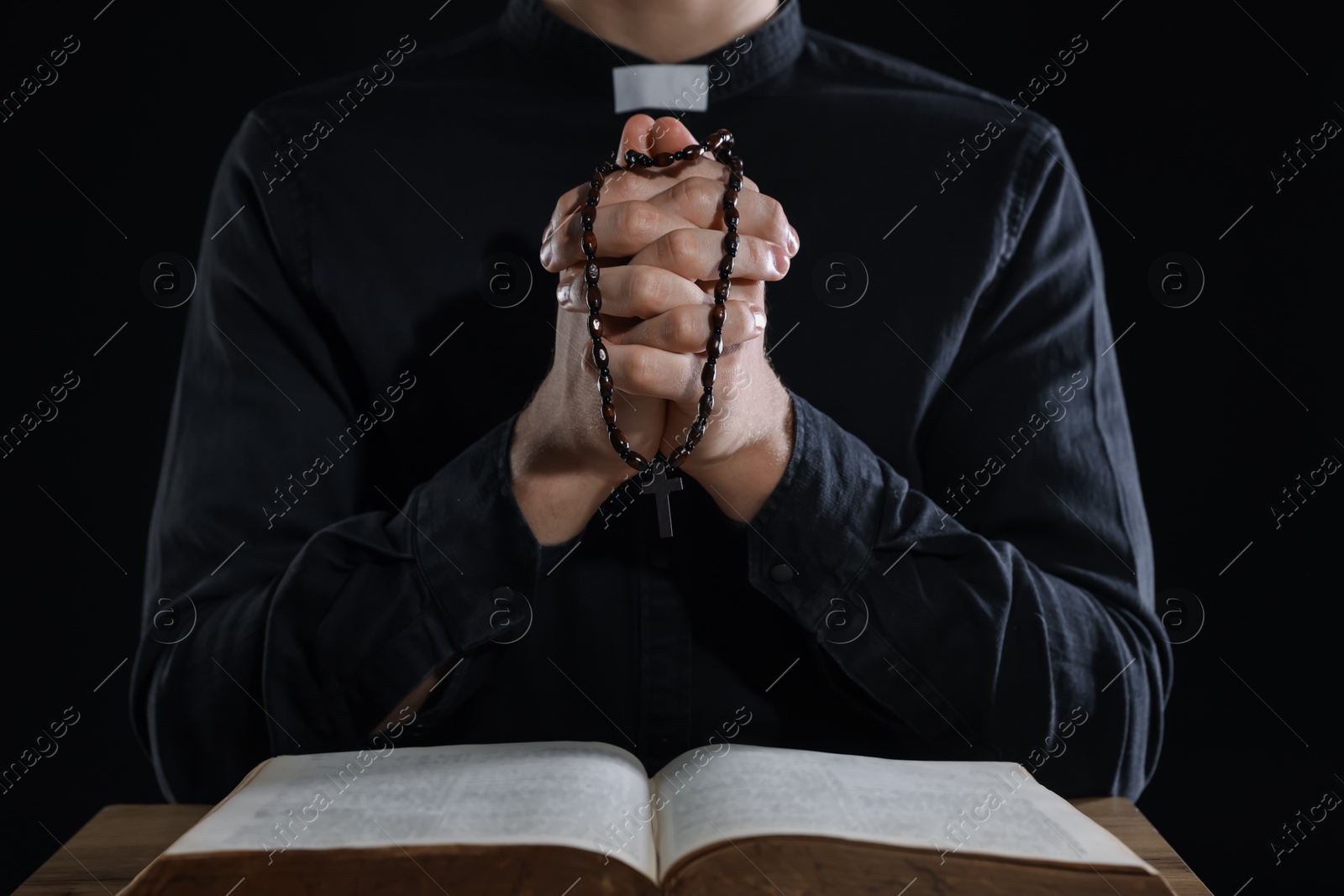 Photo of Priest with rosary and Holy Bible praying at table on black background, closeup