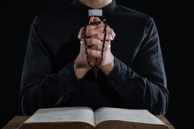 Photo of Priest with rosary and Holy Bible praying at table on black background, closeup