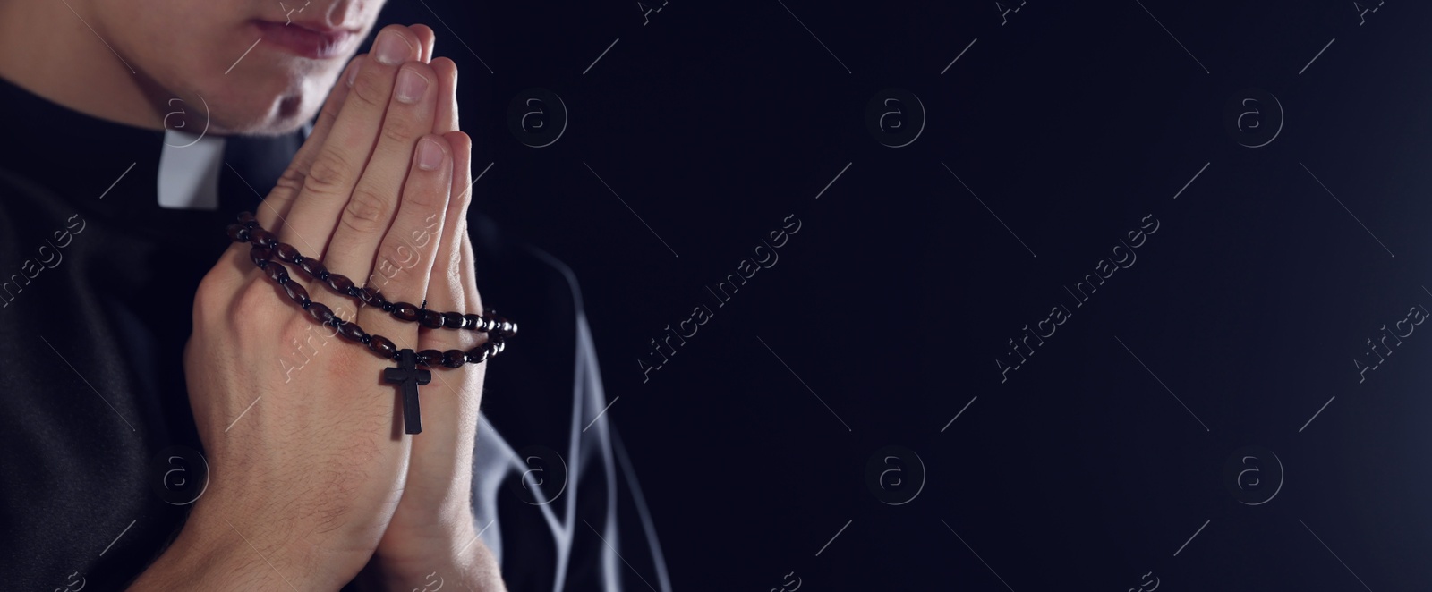 Photo of Priest with rosary praying on dark background, closeup. Space for text