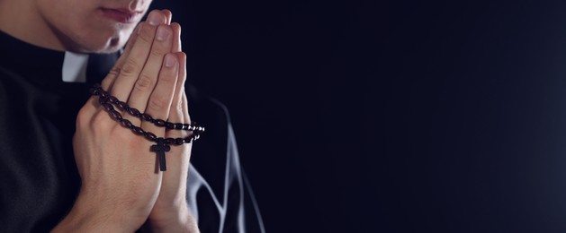 Photo of Priest with rosary praying on dark background, closeup. Space for text