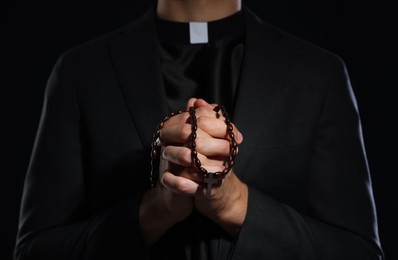 Photo of Priest with rosary praying on black background, closeup