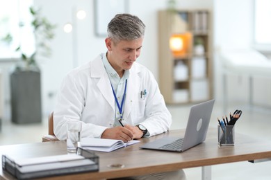 Photo of Telemedicine. Doctor making notes while having video call with patient via laptop indoors
