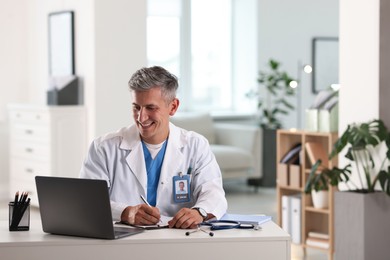 Photo of Telemedicine. Doctor making notes while having video call with patient via laptop indoors
