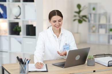 Photo of Telemedicine. Doctor making notes while having video call with patient via laptop indoors