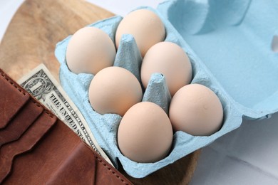 Photo of Raw chicken eggs in egg carton and wallet with dollar banknote on white tiled table, closeup
