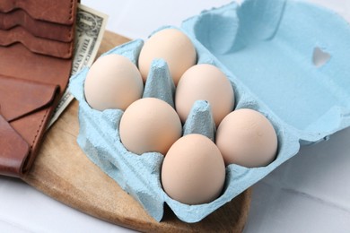 Photo of Raw chicken eggs in egg carton and wallet with dollar banknote on white tiled table, closeup
