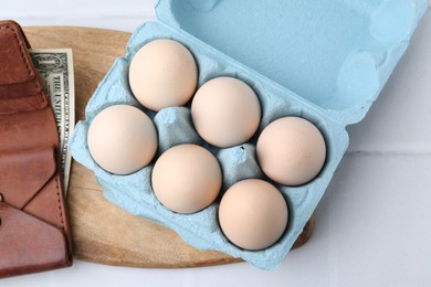 Photo of Raw chicken eggs in egg carton and wallet with dollar banknote on white tiled table, flat lay